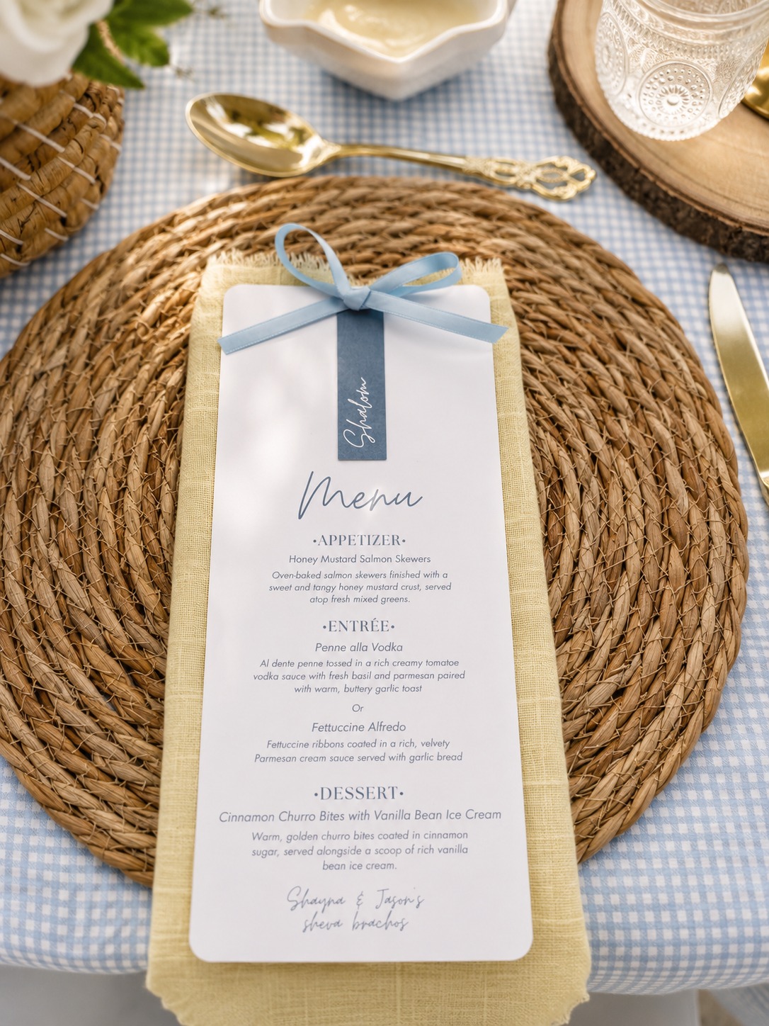A hand-designed shevabrachos menu tied with a blue ribbon on a rattan placemat and gingham linens